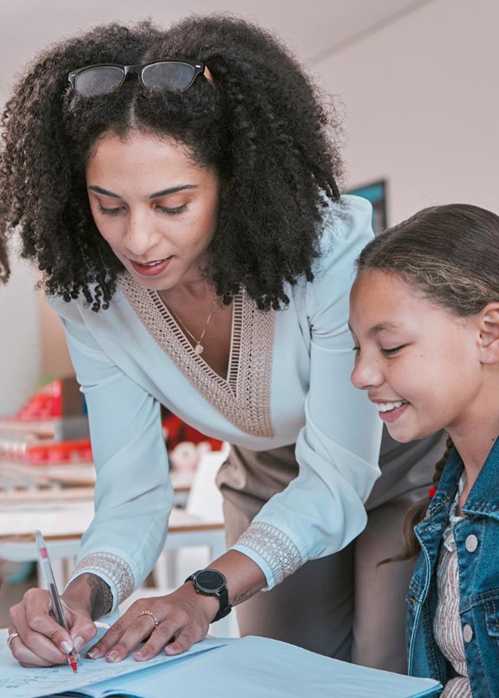 Female teacher in classroom with children helping girl student with schoolwork and writing in book diversity in education educator reading kids notebook and group learning together for assessment
